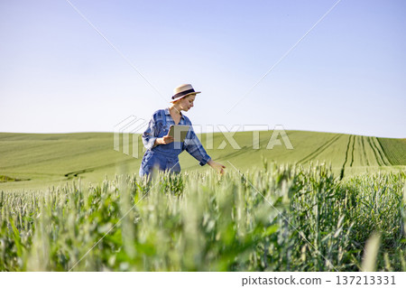 A woman in overalls and a straw hat uses a tablet to monitor the growth of a lush green wheat field under a clear blue sky 137213331