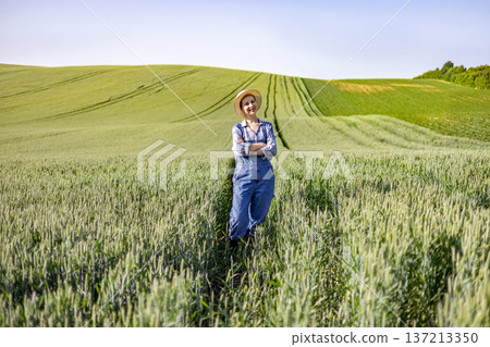 A young woman wearing overalls and a straw hat stands confidently in a vast field of ripening wheat under a clear blue sky A young woman wearing overalls and a straw hat stands confidently in a vast field of ripening wheat under a clear blue sky 137213350