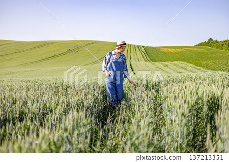 A young woman wearing overalls and a hat walks through a vast green wheat field under a clear blue sky A young woman wearing overalls and a hat walks through a vast green wheat field under a clear blue sky 137213351