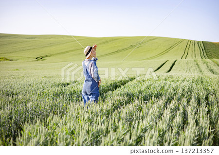 A young woman wearing denim overalls and a straw hat stands in a vast green wheat field under a clear blue sky, shielding her eyes from the sun A young woman wearing denim overalls and a straw hat stands in a vast green wheat field under a clear blue sky, shielding her eyes from the sun 137213357