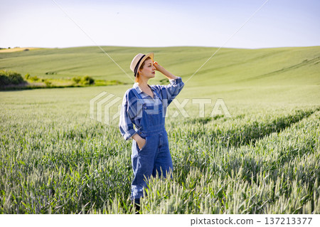 A woman wearing overalls and a hat stands in a vast green wheat field under a clear blue sky, shielding her eyes from the sun A woman wearing overalls and a hat stands in a vast green wheat field under a clear blue sky, shielding her eyes from the sun 137213377