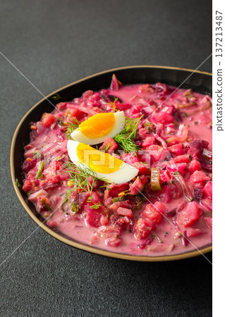 Delicious cold beet soup is garnished with boiled eggs and fresh herbs in a simple black bowl. The vibrant colors and textures create an inviting dish perfect for summer meals 137213487