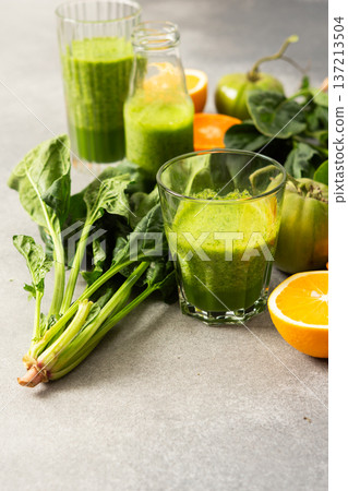 Fresh juice is made using spinach, chard, and broccoli along with oranges and tangerines. A glass of green juice sits by fresh ingredients on a clean kitchen counter 137213504