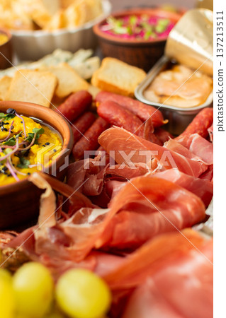 A vibrant charcuterie board showcases a variety of cheese, sausages, hummus, nuts, and sandwiches. Friends gather at a sunny outdoor picnic, enjoying delicious food together A vibrant charcuterie board showcases a variety of cheese, sausages, hummus, nuts, and sandwiches. Friends gather at a sunny outdoor picnic, enjoying delicious food together 137213511