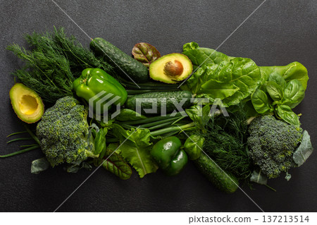 Various green vegetables such as broccoli, cucumbers, green peppers, and lettuce, fresh herbs like basil and dill, along with ripe avocado are artfully arranged on a dark background, healthy meal 137213514