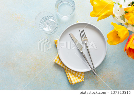 A fresh spring table setting features a plain plate, silver utensils, and a bright yellow napkin. Colorful flowers in bloom add a lively touch, creating a welcoming atmosphere for meals 137213515