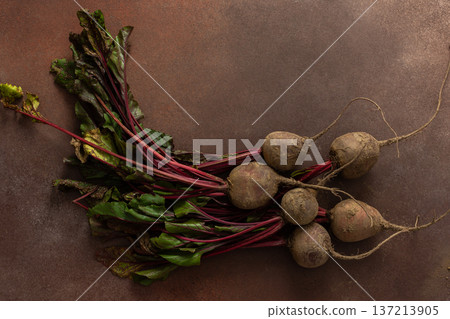 Beets with green leaves rest on a brown surface in a kitchen. The setting is warm and hints at fresh produce ready for cooking or eating Beets with green leaves rest on a brown surface in a kitchen. The setting is warm and hints at fresh produce ready for cooking or eating 137213905