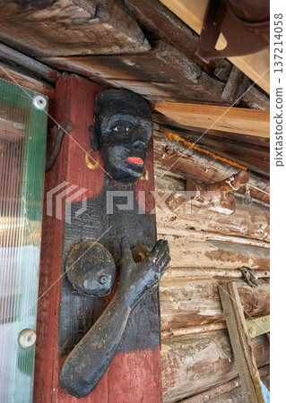 Wooden sculpture of black woman with golden earrings as decorative element on traditional wooden building. Ethnic art for rustic decor. Wooden sculpture of black woman with golden earrings as decorative element on traditional wooden building. Ethnic art for rustic decor. 137214058