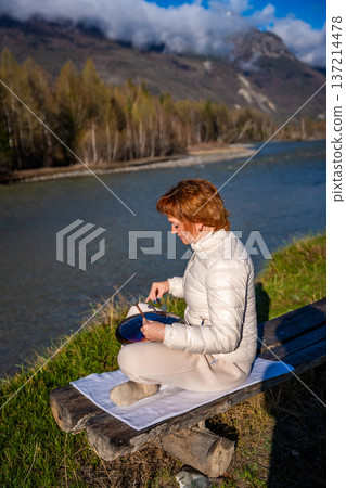 Woman playing a steel tongue drum in nature with a mountain river and Altai landscape in the background. Concept of harmony, mindfulness, and connection with nature 137214478