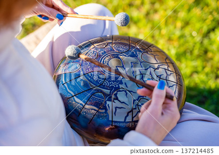 Close-up of hands playing a steel tongue drum with mallets against the backdrop of Altai mountains and river. Concept of sacred sound, touch of nature, and mindful musical ritual Close-up of hands playing a steel tongue drum with mallets against the backdrop of Altai mountains and river. Concept of sacred sound, touch of nature, and mindful musical ritual 137214485