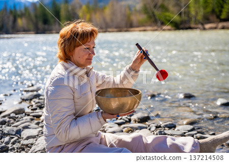 Woman performing a sound ritual with a Tibetan singing bowl by the Altai river. Concept of feminine energy, sacred vibration, and shamanic connection to the Earth Woman performing a sound ritual with a Tibetan singing bowl by the Altai river. Concept of feminine energy, sacred vibration, and shamanic connection to the Earth 137214508