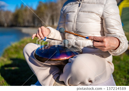 Close-up of hands playing a steel tongue drum with mallets against the backdrop of Altai mountains and river. Concept of sacred sound, touch of nature, and mindful musical ritual Close-up of hands playing a steel tongue drum with mallets against the backdrop of Altai mountains and river. Concept of sacred sound, touch of nature, and mindful musical ritual 137214589