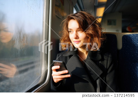 Woman in black coat traveling by train, gazing at smartphone, illuminated by soft sunlight, creating a serene atmosphere with reflections on the window 137214595
