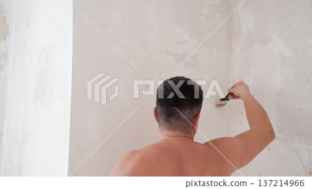 Shirtless man applying primer to interior wall with paintbrush during apartment renovation process 137214966
