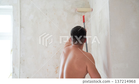 Shirtless man painting upper corner of wall with roller on extension pole during apartment renovation Shirtless man painting upper corner of wall with roller on extension pole during apartment renovation 137214968