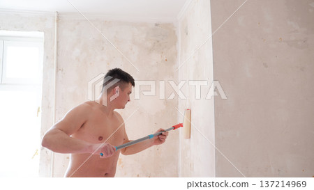 Shirtless man painting interior wall with roller on extension pole in bright unfinished room Shirtless man painting interior wall with roller on extension pole in bright unfinished room 137214969