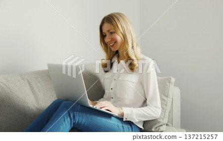 Happy modern woman working with laptop, freelancer girl sitting on a couch in the living room Happy modern woman working with laptop, freelancer girl sitting on a couch in the living room 137215257