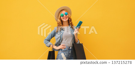 Beautiful happy young woman holding phone with black shopping bag, summer hat on yellow background 137215278