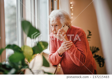 Old woman standing by window, making phone call. Old woman standing by window, making phone call. 137215483