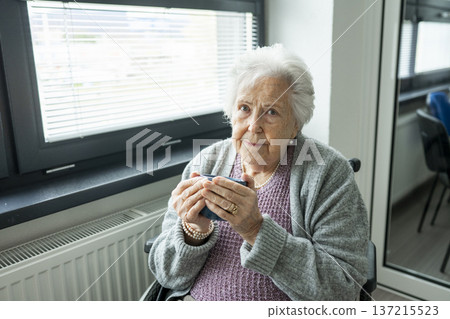 Senior woman in wheelchair holding warm cup, looking at camera. Senior woman in wheelchair holding warm cup, looking at camera. 137215523
