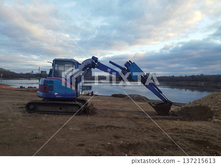 A small, blue and magenta excavator sits on muddy, brown earth by a still body of water, set against a wide, cloudy sky during twilight or a dull day 137215613