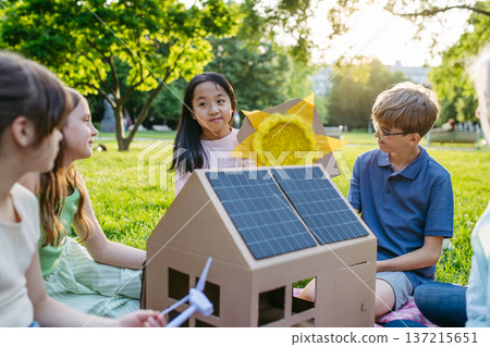 Children learning about renewable energy and solar panels during sustainable education class outdoors, using cardboard model of house wit solar panel on roof. Children learning about renewable energy and solar panels during sustainable education class outdoors, using cardboard model of house wit solar panel on roof. 137215651