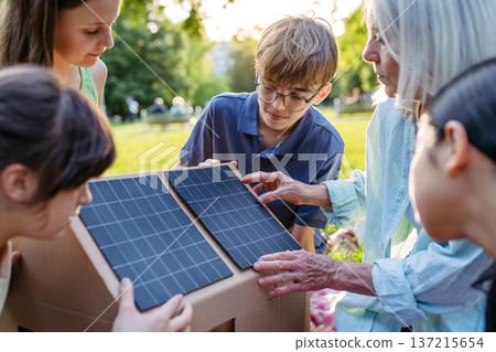 Children learning about renewable energy and solar panels during sustainable education class outdoors, using cardboard model of house wit solar panel on roof. Children learning about renewable energy and solar panels during sustainable education class outdoors, using cardboard model of house wit solar panel on roof. 137215654