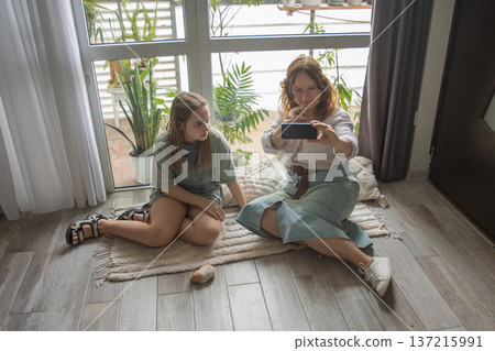 A mother and her daughter sit on a rug with hamster in a well-lit room. They are focused on a phone A mother and her daughter sit on a rug with hamster in a well-lit room. They are focused on a phone 137215991