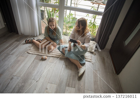 Mother and daughter sitting on a rug in a modern room while watching a video on a phone Mother and daughter sitting on a rug in a modern room while watching a video on a phone 137216011