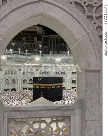 Night view of the Kaaba in Mecca framed by an ornate marble arch with pilgrims around tawaf 137216173