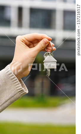 Vertical Closeup Woman Holds House Key, Apartment Building Stands In Background. Homeownership, Real Estate Deal, Purchase, Moving, Investment. Sale Buy Apartment, Mortgage, First Homeowner, New Home 137216517