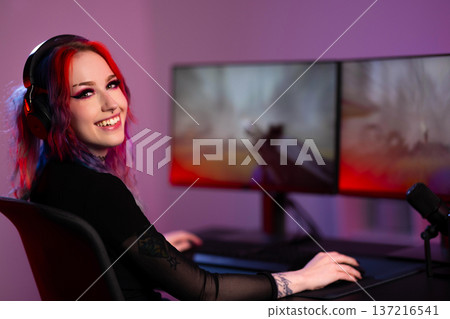 Smiling young woman with vibrant hair streaming gameplay with dual monitors Smiling young woman with vibrant hair streaming gameplay with dual monitors 137216541