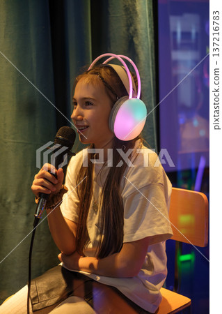 A girl in luminous headphones sings into a microphone at a karaoke club. The child gives an interview in a TV and radio company A girl in luminous headphones sings into a microphone at a karaoke club. The child gives an interview in a TV and radio company 137216783