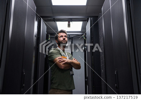 Data center technician standing between server racks with arms crossed looking up while monitoring equipment Data center technician standing between server racks with arms crossed looking up while monitoring equipment 137217519