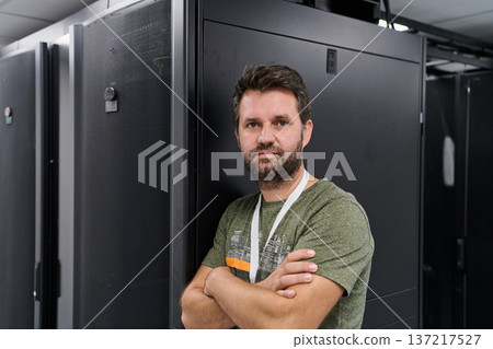 Data center technician standing with arms crossed beside server racks in modern IT infrastructure room 137217527