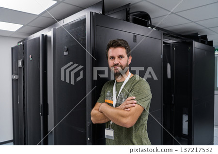 IT technician standing confidently in a data center next to server racks wearing an ID badge IT technician standing confidently in a data center next to server racks wearing an ID badge 137217532