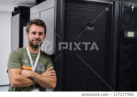 IT technician standing by server rack in a modern data center with crossed arms and badge 137217560