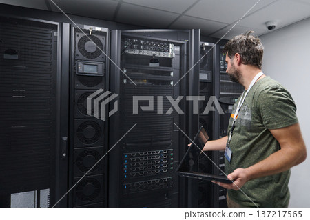 IT technician inspecting servers in a data center with a laptop, performing maintenance and network checks IT technician inspecting servers in a data center with a laptop, performing maintenance and network checks 137217565