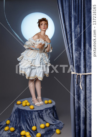 Young woman in vintage dress standing on pedestal with lemons in studio with blue curtain Young woman in vintage dress standing on pedestal with lemons in studio with blue curtain 137218165