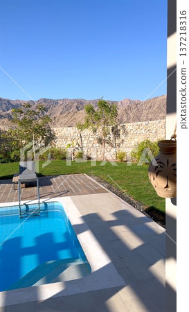 Sunlit Pool Terrace With Mountain Backdrop And Loungers Beside Clear Blue Water, Palm Trees And Manicured Lawn Under Bright Sky Conveying Relaxed Staycation Mood 137218316