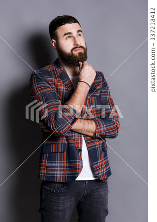 Pensive young bearded hipster looking upwards. Handsome relaxed guy in checkered jacket dreaming about something, lost in thoughts, gray studio background Pensive young bearded hipster looking upwards. Handsome relaxed guy in checkered jacket dreaming about something, lost in thoughts, gray studio background 137218412