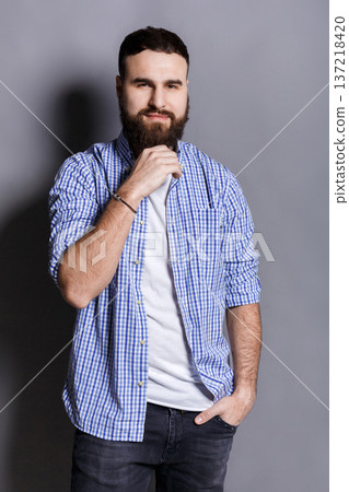 Pensive bearded man posing with hand on chin. Portrait of young confident guy, looking at camera, gray studio background Pensive bearded man posing with hand on chin. Portrait of young confident guy, looking at camera, gray studio background 137218420