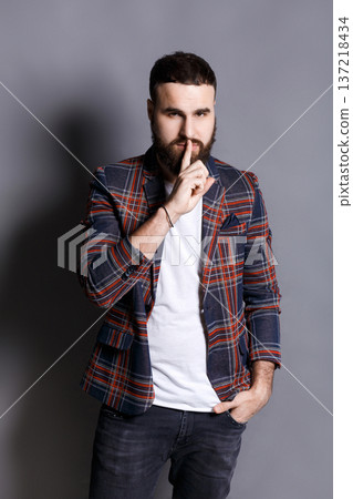 Bearded man shows hush be quiet. Handsome hipster holding finger at his lips, gray studio background, copy space. Secret, privacy concept 137218434