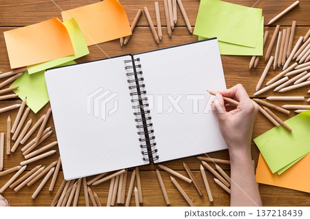 A person's hand holds a pencil over an open sketchbook surrounded by colored pencils and sticky notes on a wooden table. The setting is organized for creative brainstorming. 137218439