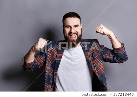 Excited bearded man very glad to success. Young cheerful guy in checkered jacket smiling at camera and gesturing with hands, gray studio background Excited bearded man very glad to success. Young cheerful guy in checkered jacket smiling at camera and gesturing with hands, gray studio background 137218446