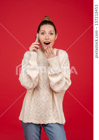 Young woman on red background speaking on the phone and looking shocked 137218453