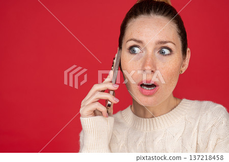 Young woman on red background speaking on the phone and looking astonished 137218458