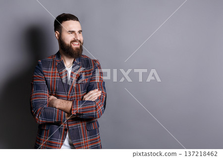 Handsome confident bearded man portrait. Young hipster in checkered jacket with crossed hands smiling away, gray studio background, copy space Handsome confident bearded man portrait. Young hipster in checkered jacket with crossed hands smiling away, gray studio background, copy space 137218462