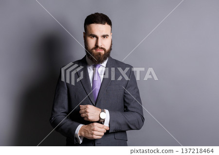 Handsome young bearded man in suit adjusting sleeves. Confident stylish businessman ready for meeting, copy space, gray studio background Handsome young bearded man in suit adjusting sleeves. Confident stylish businessman ready for meeting, copy space, gray studio background 137218464