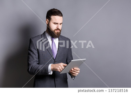 Young bearded businessman using tablet computer, copy space. Confident man in suit reading news or contract, chatting, online banking, surfing the net. Young bearded businessman using tablet computer, copy space. Confident man in suit reading news or contract, chatting, online banking, surfing the net. 137218516